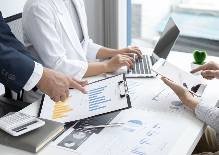 person pointing to a bar graph while two other people take notes on their laptop and ipad